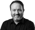 Black and white headshot of Zane Shewalter, smiling and looking at the camera. He has a beard and is wearing a black collared shirt.