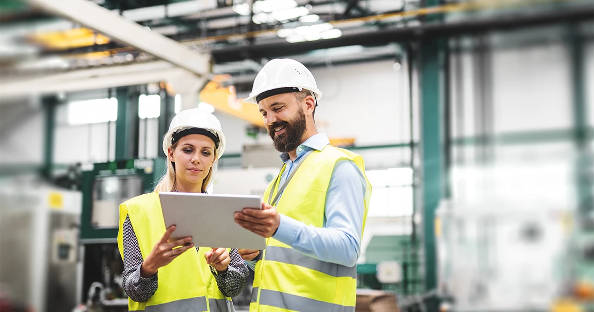 Two manufacturing professionals, a man and a woman, wearing hard hats and high-visibility vests, review data on a tablet in a factory setting. The man smiles as they discuss production.