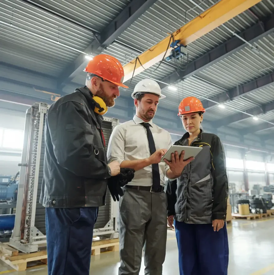 Three manufacturing professionals in hard hats, two men and one woman, review a tablet together on a factory floor. Focus on shop floor operations.