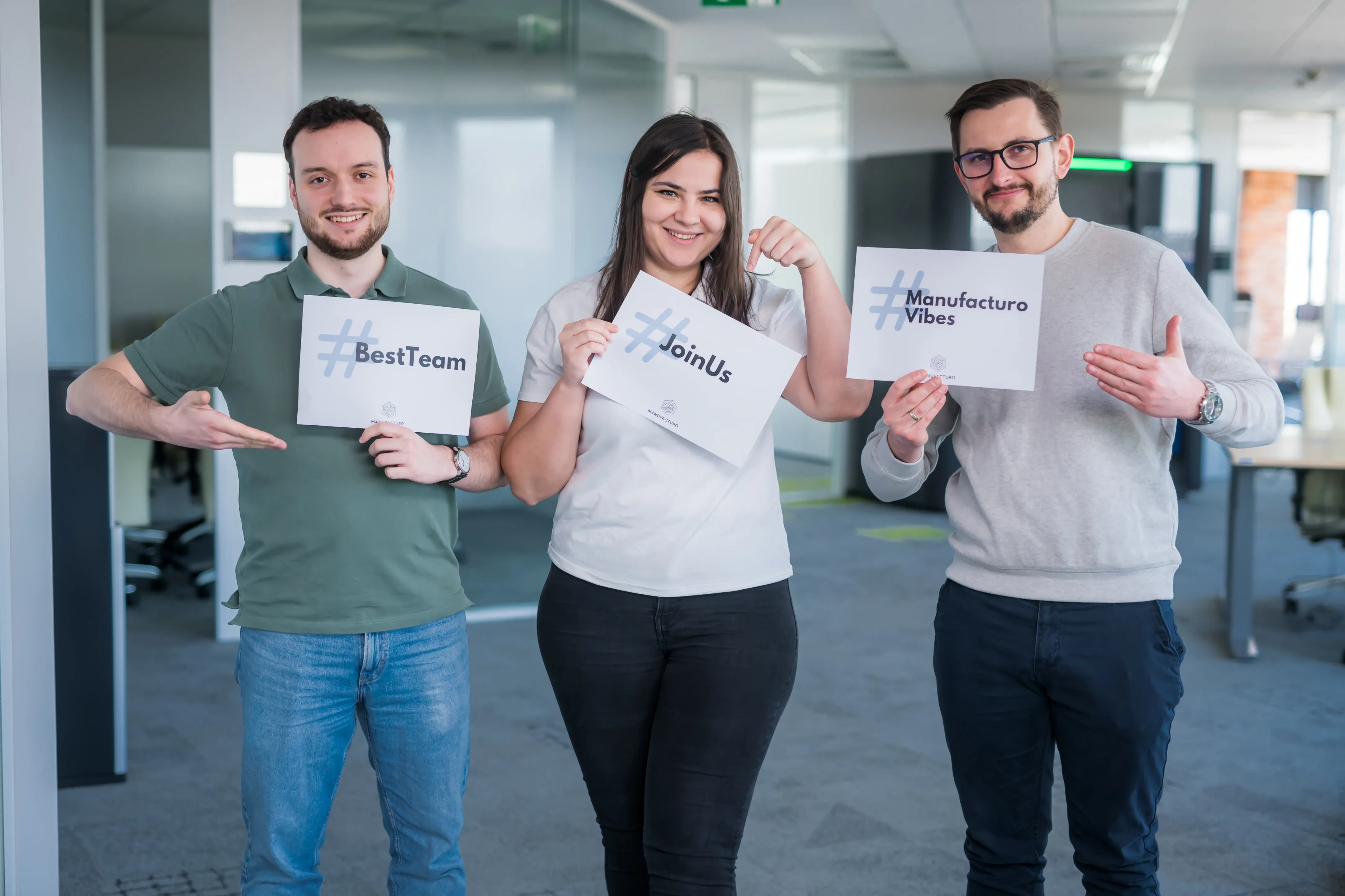 Two men smiling in an office setting. The man on the left wears a grey hoodie with the Manufacturo logo. The man on the right wears glasses and holds two signs: #Manufacturo Vibes and #Challenge Accepted. Modern office background.